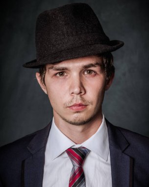 Siyah şapkalı, beyaz gömlekli, siyah ceketli, stüdyoda koyu renk arka planda kırmızı kravatlı genç bir adam portresi.