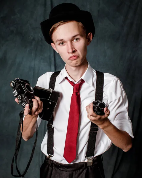 Un jeune homme portant une chemise blanche, un chapeau noir et une cravate rouge avec des bretelles tient une caméra démontée dans ses mains et regarde la caméra avec surprise. — Photo