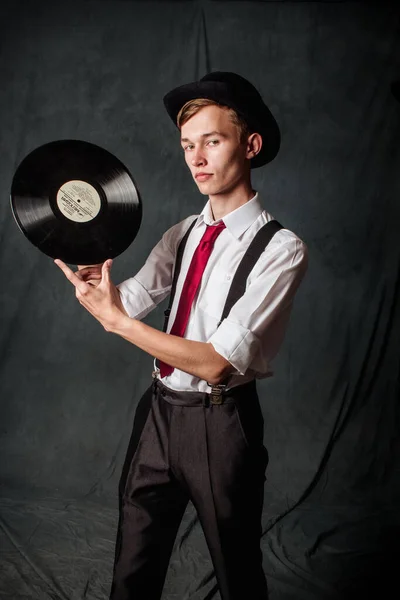 Un gars en chemise blanche, cravate rouge, chapeau noir, pantalon noir et bretelles tient une plaque de musique regardant dans la caméra sur un fond sombre dans le studio. — Photo