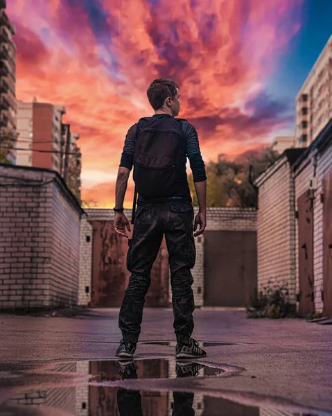 Ein junger Mann in dunkler Kleidung mit Rucksack steht mit dem Rücken zwischen Garagen vor dem Hintergrund von Hochhäusern und dem untergehenden roten Himmel und spiegelt sich in einer Pfütze. lizenzfreie Stockfotos
