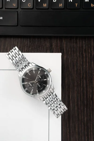 Наручные часы с серебряным браслетом и черным циферблатом лежат на книге рядом с клавиатурой на столе из темного дерева Лицензионные Стоковые Фото