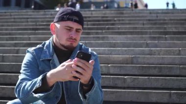 Genç adam basamaklarda oturur ve telefonu mavi gökyüzüne tutar.