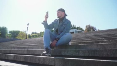 Genç adam basamaklarda oturur ve telefonu mavi gökyüzüne tutar.