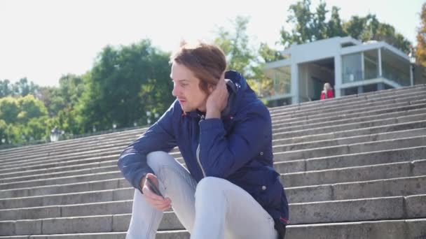 El tipo se sienta en los escalones y se comunica emocionalmente en el teléfono con chaqueta azul — Vídeo de stock