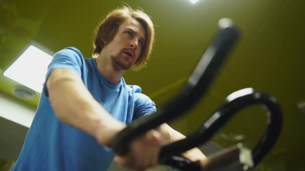 Ο κοκκινομάλλης με το μπλε μπλουζάκι οδηγεί ένα ποδήλατο στο γυμναστήριο. κοντινό πλάνο ενός χεριού — Αρχείο Βίντεο