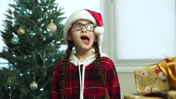 Menina sorri no chapéu de Papai Noel. Árvore de Natal no fundo. — Vídeo de Stock