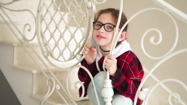 Gözlüklü küçük kız. Merdivenlere oturur ve kafasını parmaklıkların arasına sokar. — Stok video