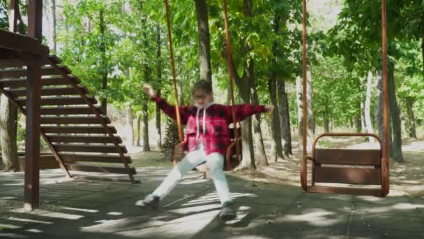Klein meisje in bril en een rode trui is rijden een swing.green pretpark — Stockvideo