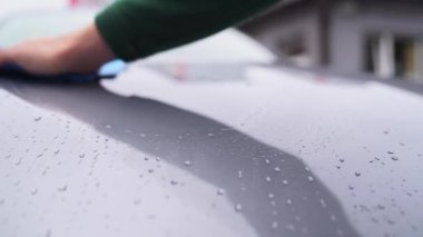 Bir adam gri bir arabanın ıslak kaputunu mavi bir paçavra makinesiyle siliyor.