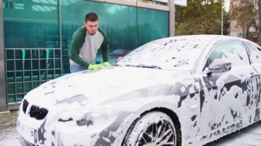 Genç adam arabasını yıkıyor. Gri bir arabanın yan camını ovar. 