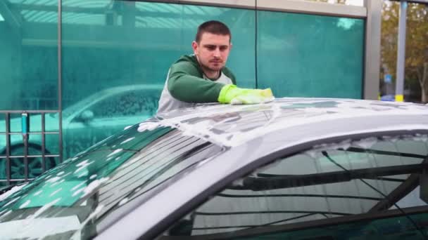 Facet myje swój szary samochód w gumowych rękawiczkach na myjce serwisowej.dużo piany na dachu samochodu — Wideo stockowe
