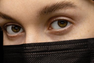 Siyah tıbbi maskeli genç bir kadının yüzünün bir parçası. Sakin kahverengi gözler.