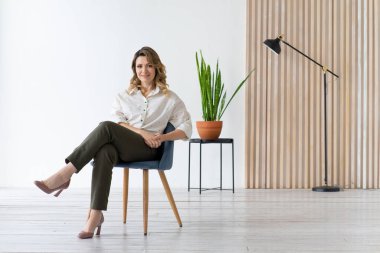 Mutlu orta yaşlı kadın sandalyeye oturur. Minimalist iç mekan. Kopyalama alanı olan beyaz arkaplan.