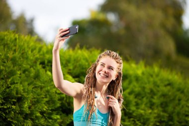 Kafasında rasta olan genç bir kadın parkta telefonda selfie çekiyor..