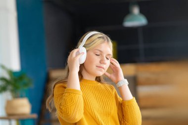 Sarı kazaklı genç kız müzik dinlemek için kablosuz beyaz kulaklık kullanıyor. Kopya alanı olan ev içi arkaplan.