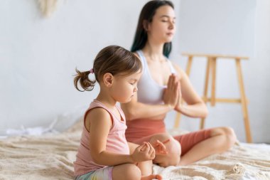 Genç anne küçük kızıyla kanepede yoga yapıyor. Lotus pozisyonu meditasyonu. Padmasana egzersizi.