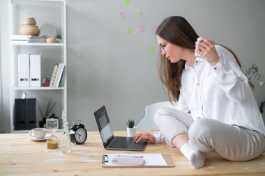 Kadın elinde kupayla masada oturuyor ve bilgisayarını kullanıyor..