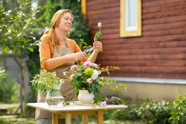 Happy middle-aged red-haired woman florist makes a handmade bouquet in her garden. Cuts the stem of a flower with a pruner.
