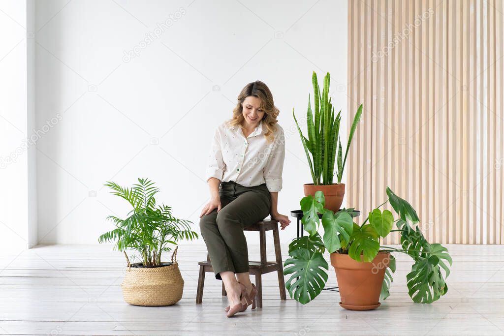 Happy middle-aged woman sits on chair next to houseplants. Minimalistic interior, japandi.