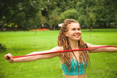 Spor kıyafetleri içinde kırmızı elastik bantla parkta antrenman yapan gülümseyen genç bir kadın.. 