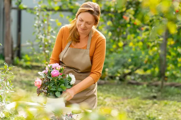 A happy middle-aged woman in an orange sweatshirt and beige apron makes a handmade bouquet in her garden. Home hobby in early fall. Blure foreground.