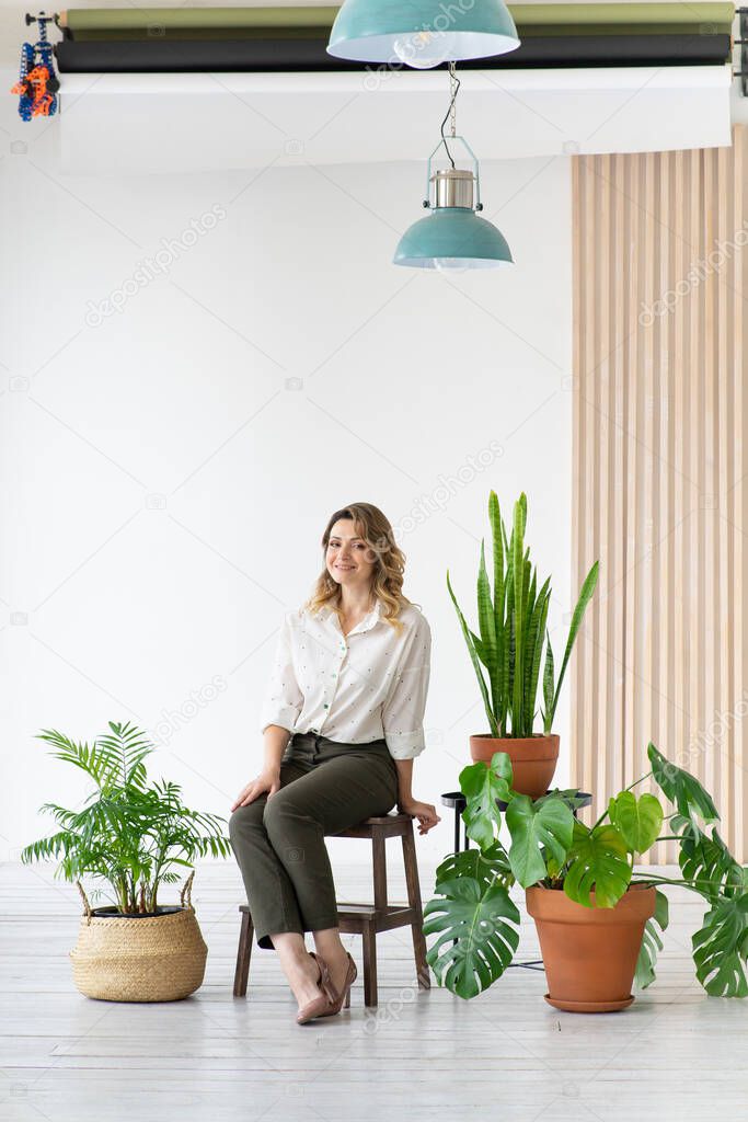 Happy middle-aged woman sits on chair next to houseplants. Minimalistic interior, japandi. White background with copy space.