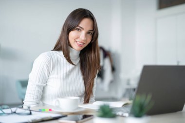 Beyaz kazaklı, gülümseyen bir kadın parlak odada dizüstü bilgisayarla oturuyor. Merkez Ofis.