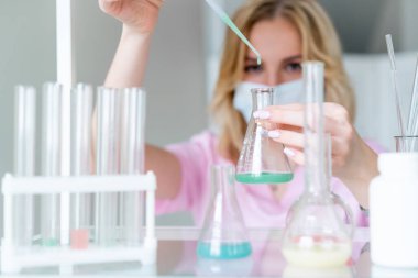 Pembe önlüklü kadın mikrobiyolog laboratuarda araştırma yapıyor, yeşil sıvıyı pipetle birlikte mavi önplanlı, renkli sıvıyla şişenin içine bırakıyor..