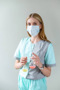 Tıbbi maskeli genç kadın bilim adamı araştırma yapıyor ve iki adet düz tabanlı kırmızı ve sarı sıvı şişeyi elinde tutarak tıp alanında geniş bir keşif yapıyor..
