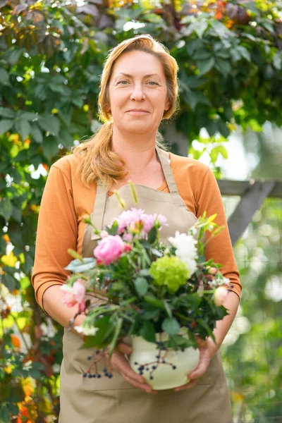 Gardening, floristics and middle aged people concept - portrait of happy redhead woman in beige garden apron with bunch of flower over garden background. Blurred foregriound.