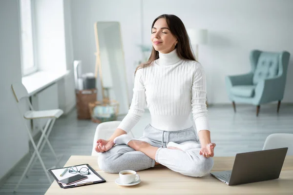 Padmasana Yoga 'da laptopun yanında gözleri kapalı oturan genç bir kadın ev ofisine poz veriyor..
