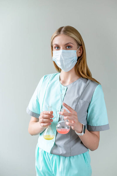 Young woman scientist in medical mask conducts research and makes broad discovery in field of medicine, holding two flat-bottomed flask of red and yellow liquid.