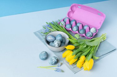 Bahar aylarında muscari çiçeği buketleri, sarı laleler, yumurtalı bir kase, gri masa peçetesinde yumurtalı mor yumurta paketleri. Metin için yer.
