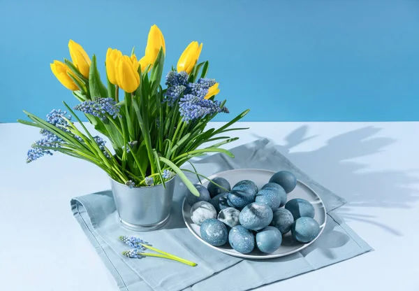 Yellow tulips, muscari in decorative bucket, plate with Easter eggs on gray kitchen napkin. A modern still life with hard shadows.