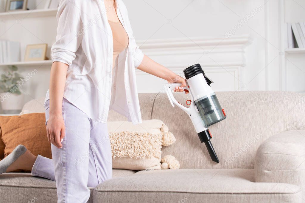 Young woman vacuums sofa with cordless handheld vacuum cleaner. House cleaning.