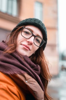 Genç bir kadının selfie 'si. Gözlüklü bir kadın kameraya bakar. Sıcak bir kışlık ceket, şapka ve atkıyla. Günlük makyaj malzemesi. Arka planda uzun turuncu bir bina var..