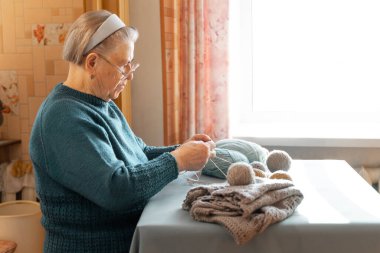 Yaşlılar. Gri saçlı bir kadın mutfak masasında otururken örgü örüyor. Görüş için gözlük takıyor. Masanın üzerinde iplik topları ve örgü bir ceket var. En sevdiği hobi.