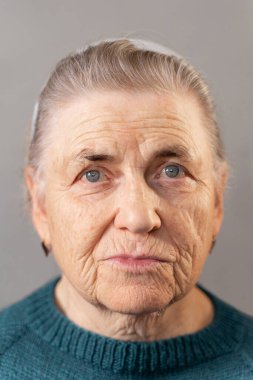 80 'li yaşlarda, kırışıklıkları derin, yaşlı bir kadının yüzü. Kameraya bak. Örgü yeşil ceketli gri saçlı yaşlı bir kadının portresi. Gri arkaplan.