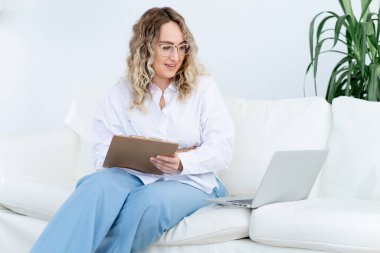 Mutlu Kadın Psikolog hastasıyla yaptığı video görüşmesi sırasında not defterine not alıyor. Minimalist ve hafif ofisindeki koltukta oturuyor. Doktorla online randevu.