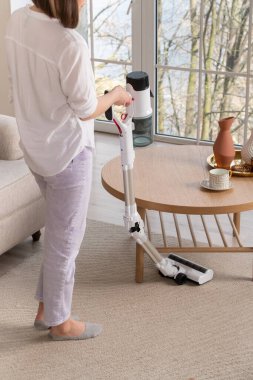Açık tişörtlü ve pantolonlu bir kadın büyük pencereli oturma odasındaki tahta masanın altını süpürüyor. Bej renkli halı. Bükme kolu fonksiyonu olan elektrikli süpürge. Elektrik süpürgesinin sapındaki toz toplayıcısı..