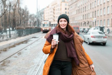 Caddenin ortasında turuncu ceketli ve bir kupa kahveyle dolaşan mutlu yüzlü bir kızıl. Kameraya bak. Şehrin bulanık görüntüsü.