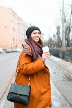 Kızıl saçlı bir kadın soğuk mevsimde gülümser ve sokakta yürür. Turuncu uzun ceket, atkı ve kalın şapka. Bir fincan sıcak kahve. Başka tarafa bakıyor. Şehrin bulanık görüntüsü.