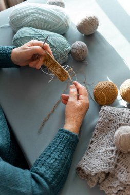 Yaşlı bir kadının elleri doğal yün iplikler ve örgü iğneleri kullanarak örüyor. Masanın üzerinde mavi bir masa örtüsü, iplik topları ve örgü bir ceket var. Ellere odaklan. Üst görünüm.