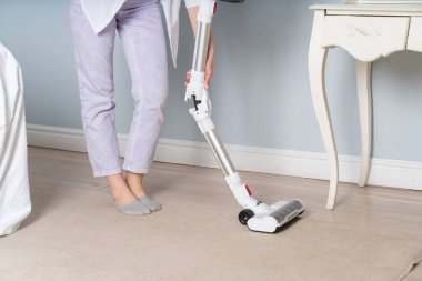 Pastel mor pantolonlu kadın bacakları. Hafif çarşaf. Ulaşılması zor yerlerde çöp toplamak için bükme işlevine sahip elektrikli süpürge. Bacaklar görünmez.. 