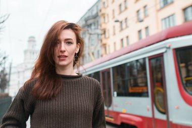 Tramvay durağında toplu taşıma bekleyen bir kadın. Gidiş gelişler, günlük kıyafetler. Kameraya bak. Modern bir tramvayın ve şehir sokağının arka planında.