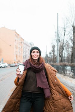 Kızıl saçlı gülümsedi ve yüzünde mutlu bir ifadeyle caddede yürüdü. Turuncu uzun bir ceket ve bir kupa kahveyle. Başka tarafa bakıyor. Şehrin bulanık görüntüsü.