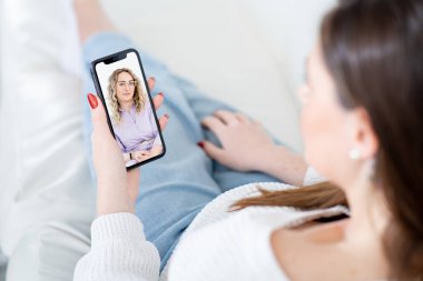 Kadın hasta, oturma odasındaki koltukta oturan cep telefonuyla video çağrısında dikkatli bayan psikolog tavsiyesi dinliyor.