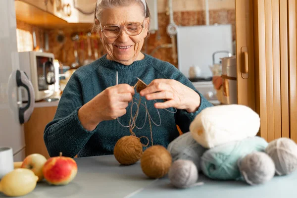 Happy elderly woman is engaged in her favorite hobby, knitting while sitting in kitchen. Glasses for vision. There are threads on table, a cup of tea, apples and a lemon. Focus on face elderly woman