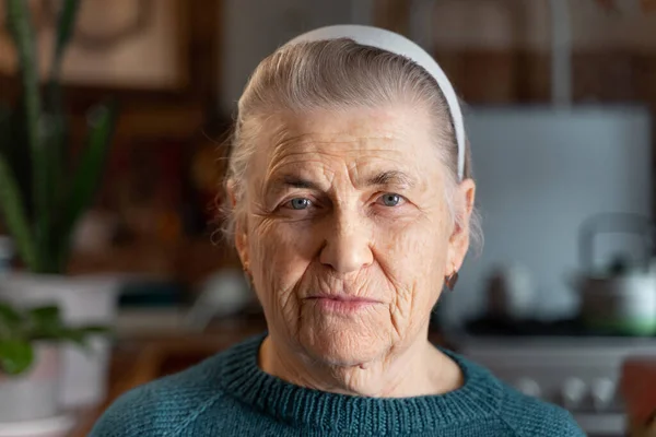 An old woman with deep wrinkles and gray hair looks at the camera. There is a headband on the head. Green knitted jacket. The concept of old age.