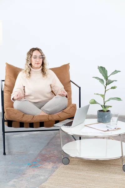 Gözlüklü sarışın bir kadın nilüfer çiçeği pozisyonunda bir sandalyede meditasyon yapıyor. Masanın üzerinde açık bir dizüstü bilgisayar ve meditasyon üzerine video dersi var. Evde dinlenme ve rahatlama..